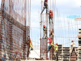 Bauarbeiter mit Schutzhelmen und Sicherheitsgurten arbeiten an einem Stahlbetongerüst auf einer Großbaustelle unter freiem Himmel