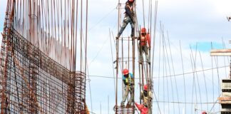 Bauarbeiter mit Schutzhelmen und Sicherheitsgurten arbeiten an einem Stahlbetongerüst auf einer Großbaustelle unter freiem Himmel