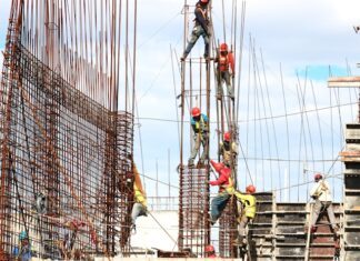 Bauarbeiter mit Schutzhelmen und Sicherheitsgurten arbeiten an einem Stahlbetongerüst auf einer Großbaustelle unter freiem Himmel
