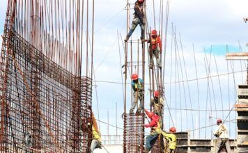 Bauarbeiter mit Schutzhelmen und Sicherheitsgurten arbeiten an einem Stahlbetongerüst auf einer Großbaustelle unter freiem Himmel