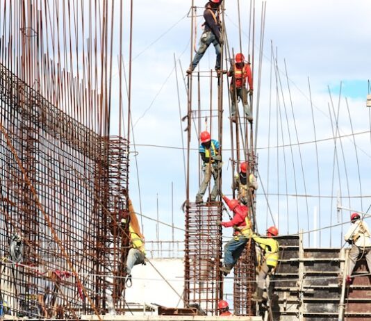 Bauarbeiter mit Schutzhelmen und Sicherheitsgurten arbeiten an einem Stahlbetongerüst auf einer Großbaustelle unter freiem Himmel