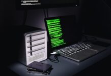 Auf einem Schreibtisch stehen ein Laptop mit grünem Programmiercode auf dem Bildschirm und ein Gehäuse für mehrere Festplatten. Daneben liegen zwei einzelne SSD-Festplatten.