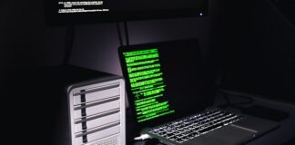 Auf einem Schreibtisch stehen ein Laptop mit grünem Programmiercode auf dem Bildschirm und ein Gehäuse für mehrere Festplatten. Daneben liegen zwei einzelne SSD-Festplatten.
