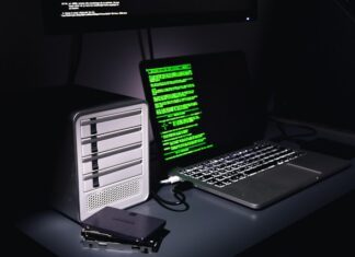 Auf einem Schreibtisch stehen ein Laptop mit grünem Programmiercode auf dem Bildschirm und ein Gehäuse für mehrere Festplatten. Daneben liegen zwei einzelne SSD-Festplatten.