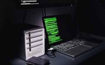 Auf einem Schreibtisch stehen ein Laptop mit grünem Programmiercode auf dem Bildschirm und ein Gehäuse für mehrere Festplatten. Daneben liegen zwei einzelne SSD-Festplatten.