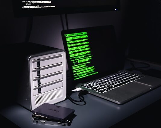 Auf einem Schreibtisch stehen ein Laptop mit grünem Programmiercode auf dem Bildschirm und ein Gehäuse für mehrere Festplatten. Daneben liegen zwei einzelne SSD-Festplatten.