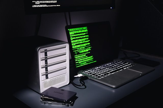 rohan-ZoXCoH7tja0-unsplash Auf einem Schreibtisch stehen ein Laptop mit grünem Programmiercode auf dem Bildschirm und ein Gehäuse für mehrere Festplatten. Daneben liegen zwei einzelne SSD-Festplatten.