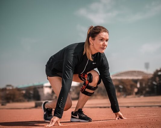 Sportlerin in Startposition auf einer Laufbahn, trägt Sportkleidung und eine Kniebandage, bereit für den Sprint.