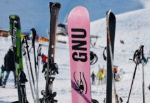 Verschiedene Skier und ein Snowboard stehen aufrecht im Schnee vor einer verschneiten Bergkulisse – Symbol für moderne und flexible Skiausrüstung, wie sie Wintersportler:innen bei Alpy.com in Nauders mieten können